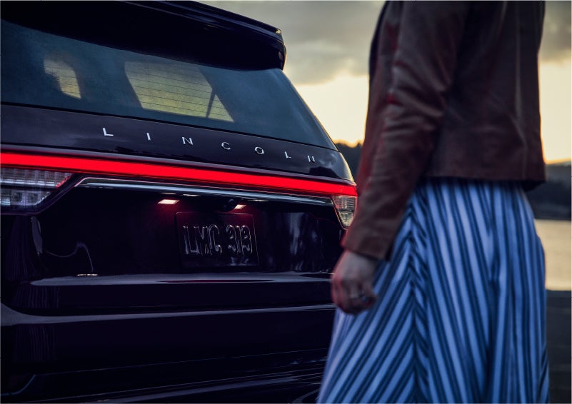A person is shown near the rear of a 2023 Lincoln Aviator® SUV as the Lincoln Embrace illuminates the rear lights | Crest Lincoln in Sterling Heights MI