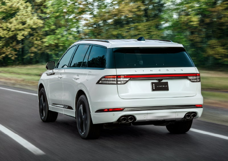 A white 2026 Lincoln Aviator® SUV is being driven away down a road lined with colorful trees. | Crest Lincoln in Sterling Heights MI