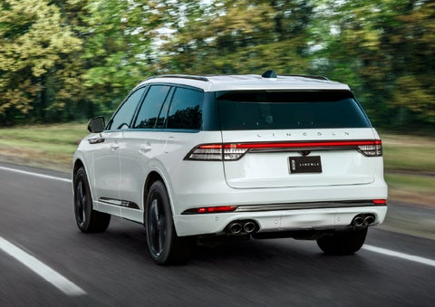 A white 2026 Lincoln Aviator® SUV is being driven away down a road lined with colorful trees. | Crest Lincoln in Sterling Heights MI