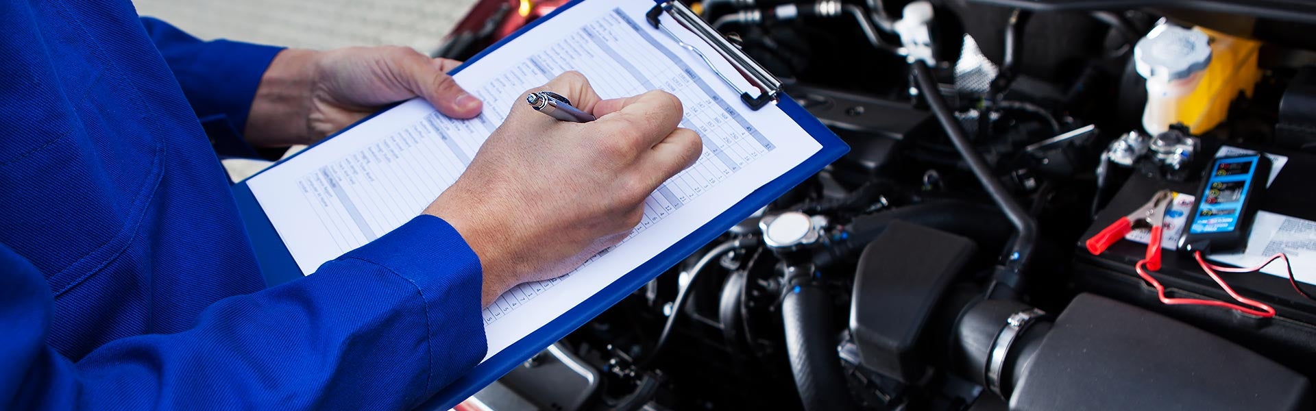 Technician in blue uniform writes on a clipboard next to an open car hood with battery and engine visible.