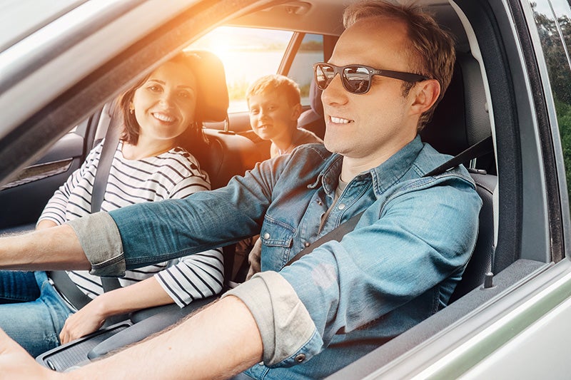 A family is smiling while driving in a car on a sunny day.
